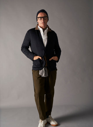 Man wearing a dark jacket, white shirt, green pants, and glasses against a gray background