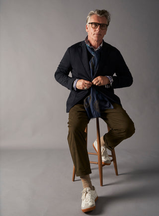 Man sitting on a stool wearing a dark jacket, green pants, and white sneakers against a gray background