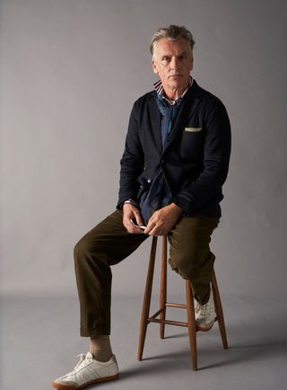 Man sitting on a stool wearing a dark jacket and brown pants against a gray background